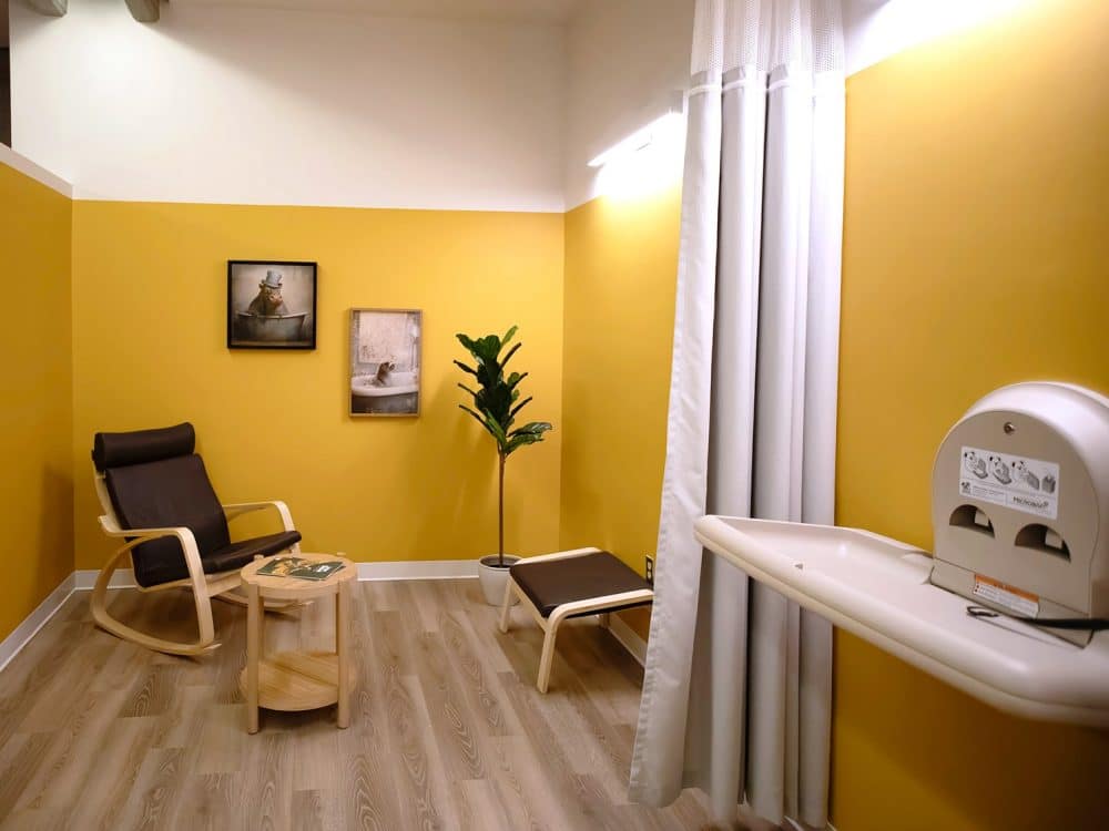 Yellow wall with two frames, an armchair on the left with a footrest on the right, a small round coffee table, a curtain and a changing table.