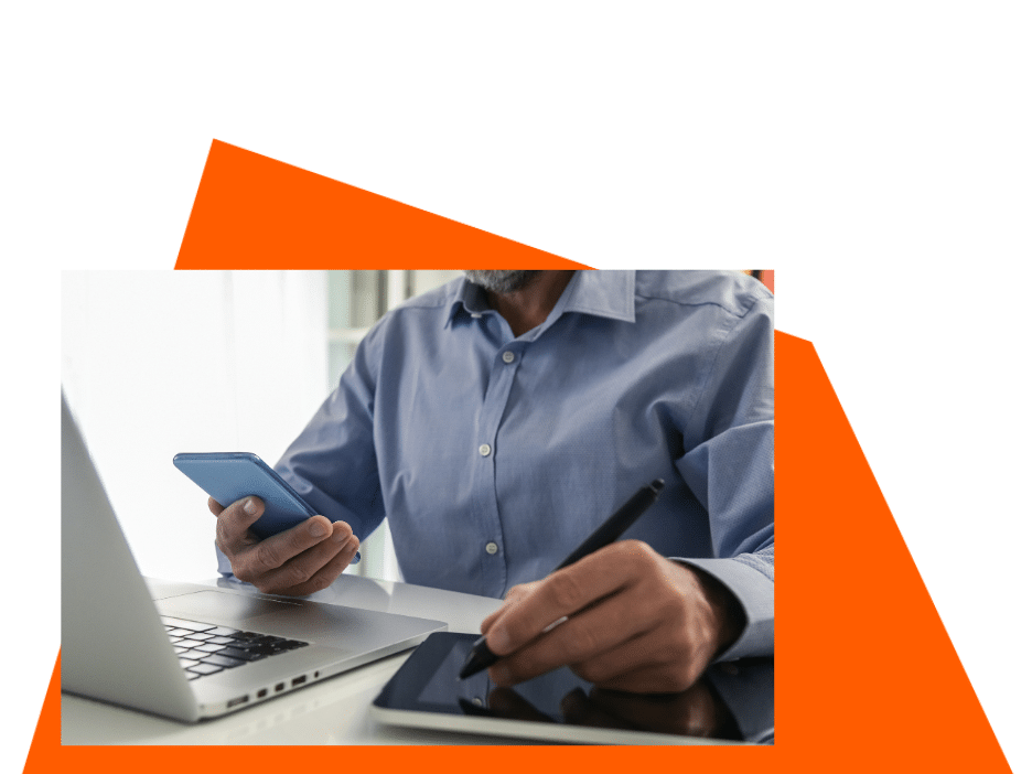 A man in front of his computer, holding his phone in one hand and a tablet on the other.