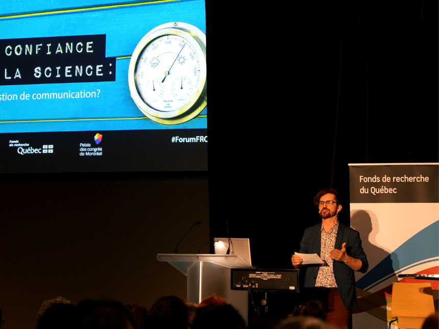 A man on stage right with a screen to his left, and a presentation on that screen.