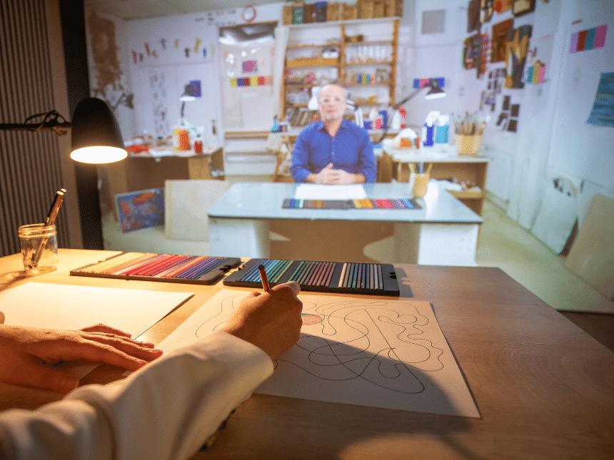 Une main posée sur une table avec des feuilles et des crayons de couleurs dessine, elle est éclairée par une petite lampe de table. Face à elle se trouve un écran géant, et dans cet écran, on y voit un artiste dans son atelier, assis à une table. 