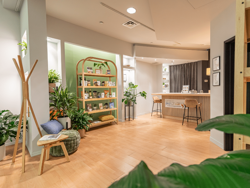 Local d'ESPACE RECHARGE avec un parquet en bois des murs beige et vert, des coussins, un ilôt de cuisine et une étagère avec des produits locaux. Le tout dans un environnement zen et cozy.