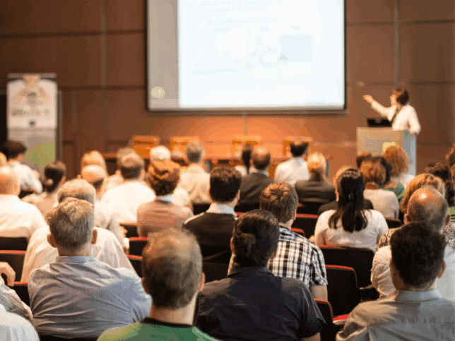 Salle d'événement avec une présentatrice, un écran et plusieurs participants assis de dos regardant la présentation.